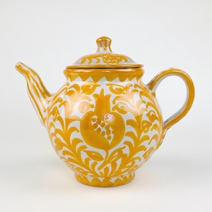 Peut inclure: Théière en céramique avec couvercle, anse et bec verseur. La théière est peinte avec un motif floral jaune et blanc, avec un motif de grenade sur le devant. La anse et le bec sont jaunes.