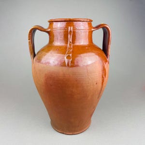 May include: A large, glazed, brown ceramic vase with two handles. The vase has a wide body that tapers towards the base and a lid. The handles are curved and attached to the upper portion of the vase. The vase is a warm, earthy tone.