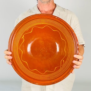 May include: A large, round, terracotta-colored ceramic bowl featuring a glossy finish. The interior surface is decorated with concentric circular lines and a single, continuous wavy line in a lighter yellow-orange hue, creating a pattern.