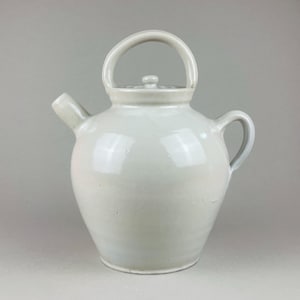 May include: A white ceramic teapot with a rounded body, a curved handle, and a spout. The teapot has a lid with a small knob and a smaller handle on the side. The teapot is a light, neutral color.