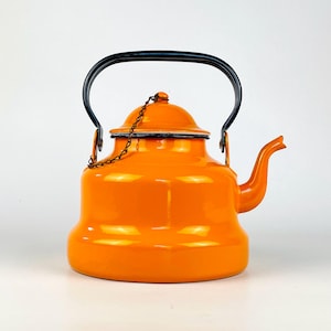 Op de afbeelding: Een feloranje geëmailleerde theeketel met een brede, ronde basis en een gebogen tuit. De ketel heeft een zwart gebogen handvat en een bijpassend oranje deksel dat met een dun metalen kettinkje aan de behuizing is bevestigd. De rand is zwart.