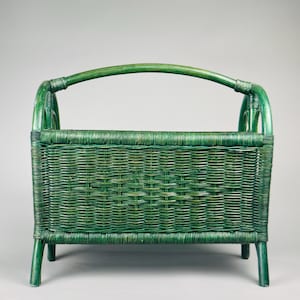 May include: A green wicker magazine rack with a curved handle. The rectangular basket is supported by four legs. The woven design is visible throughout the piece, with a consistent green color.