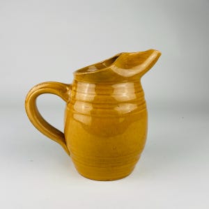 May include: A ceramic jug in a warm, golden-yellow hue, featuring a curved handle and a pouring spout. The body of the jug has horizontal ridges. The handle is arched, and the spout is angled for pouring.