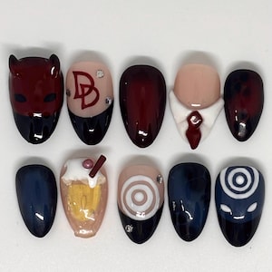 Op de afbeelding: Een set van tien kunstnagels met diverse ontwerpen, waaronder een donkerrood duivelsmasker, een huidkleurige nagel met een rood DD-logo en strass-steentjes, een effen donkerrode nagel, een witte overhemdkraag met een rode das en een blauwe roos.