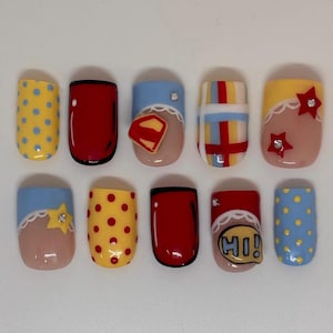Pode incluir: Um conjunto de dez unhas postiças com designs variados, incluindo amarelo com pontos azuis, vermelho sólido, azul claro com um emblema de escudo, xadrez vermelho e azul, amarelo com estrelas vermelhas, azul claro com uma estrela amarela e um balão com HI!