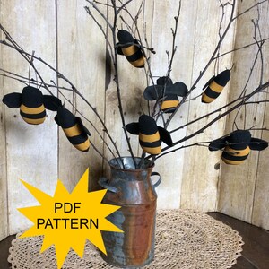 May include: A rustic arrangement featuring handmade bee ornaments hanging from bare branches in a weathered metal container. The bees are black and yellow, with black wings. The image includes a yellow starburst graphic with the text "PDF PATTERN".