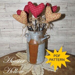 May include: A vintage metal milk churn vase displays fabric heart decorations on black sticks, tied with twine. The hearts are red and cream with floral patterns. The vase sits on a lace doily. Text on the image reads "Hoosier Hollow" and "PDF PATTERN".