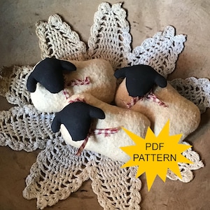 May include: Three plush sheep with black heads and red and white plaid ribbon bows. The sheep are cream-colored and rest on a cream-colored, lace doily. A yellow starburst graphic with the text "PDF PATTERN" is in the image.