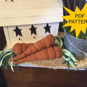 May include: Two orange fabric carrots with green and white fabric tops rest on a burlap mat. A white wooden shelf with star cutouts is in the background. A metal bucket with greenery is also visible. The image includes the text "PDF PATTERN".