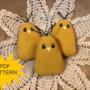 May include: Three yellow fabric chick ornaments with black eyes and beaks, each with a black string for hanging. They rest on a white, open-weave doily. A yellow starburst graphic with "PDF PATTERN" is in the lower left.