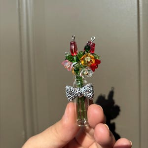 Pode incluir: Um vaso de vidro em miniatura cheio de flores de contas coloridas, incluindo flores vermelhas, amarelas e rosas. Uma fita de prata é amarrada ao redor do vaso. O vaso é segurado em uma mão.