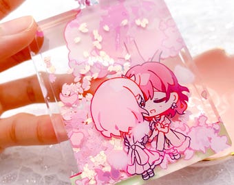 Könnte beinhalten: Ein klarer Acryl-Schlüsselanhänger mit einer rosa Kugelkette. Der Schlüsselanhänger zeigt eine rosa-weiße Illustration von zwei Anime-Figuren, die sich umarmen, umgeben von rosa Konfetti und Kirschblüten-Designs.