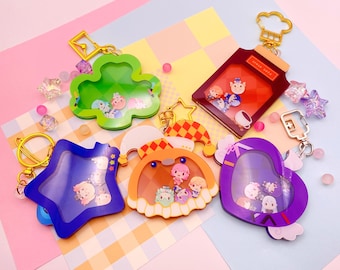 May include: A collection of colorful keychains in various shapes, including a clover, star, jar, heart, and a pumpkin-like design. Each keychain has a clear window filled with small character figures. Gold-colored key rings and charms are attached.