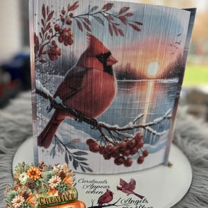 Puede incluir: Una pieza de arte de libro plegado con una escena de invierno que presenta un cardenal rojo posado en una rama con bayas rojas. El fondo muestra un paisaje nevado con una puesta de sol sobre un lago. El texto "Cardinals Appear When Angels Are Near" está escrito en la base.
