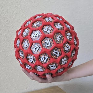 May include: A red, spherical, multi-sided die with white numbers. The die is held in a person's hand. The die has a honeycomb-like structure with numbers visible on each face. The die is likely used for tabletop games.