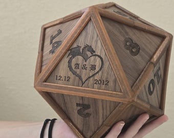 May include: A large, twenty-sided wooden die featuring dark brown wood grain. One triangular face is engraved with a dragon silhouette forming a heart around the letters A & B, with the date 12.12.2012 below. Other faces display engraved numbers.