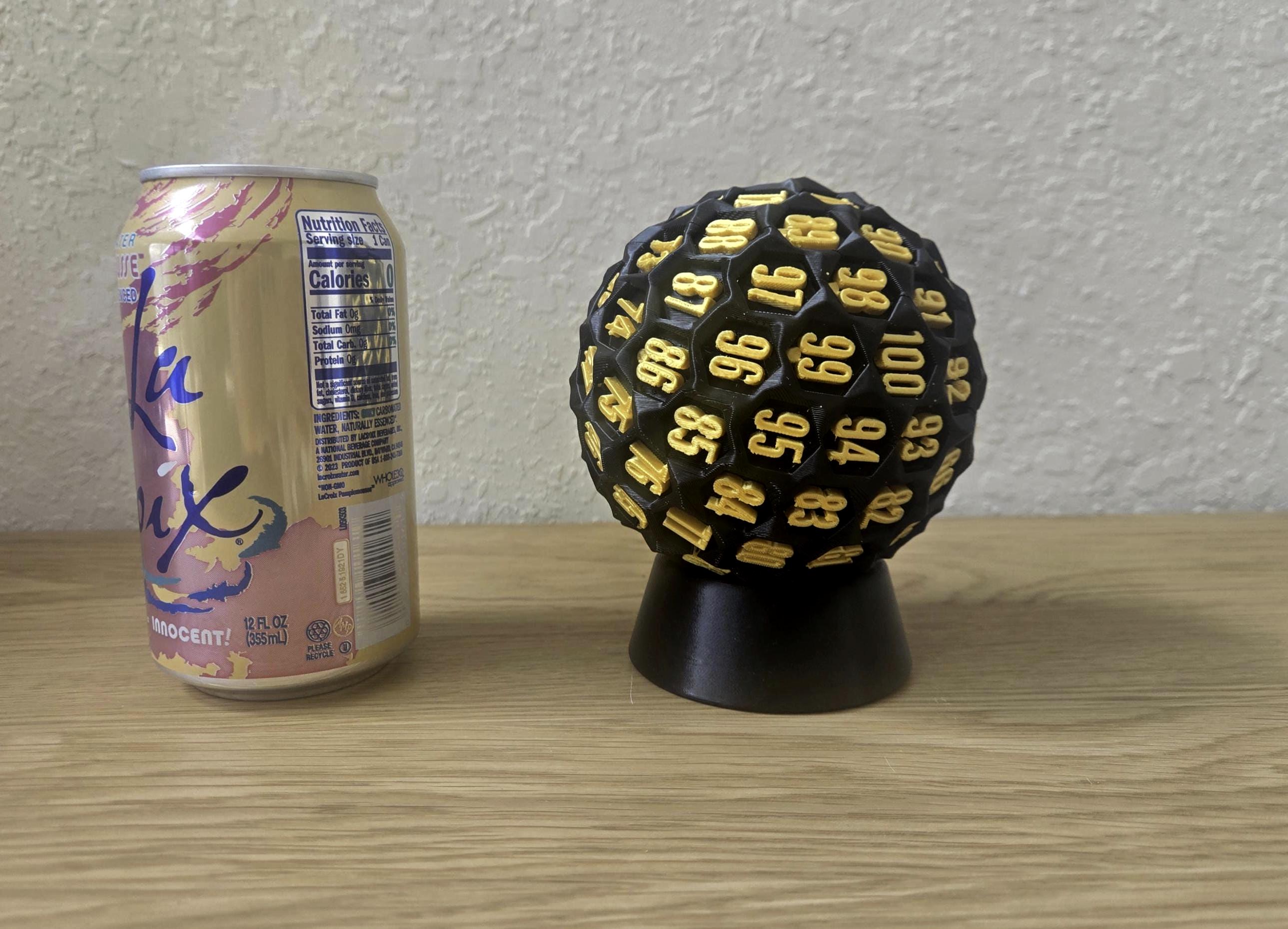 A black and gold oversized D100 dice with numbers on each face, placed on a black stand, next to a can of LaCroix water.