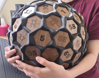 Può includere: Un grande oggetto sferico con un design geometrico. La sfera è composta da pentagoni grigio scuro ed esagoni di legno numerati. I numeri sono incisi in un carattere serif. L'oggetto è tenuto da una persona.