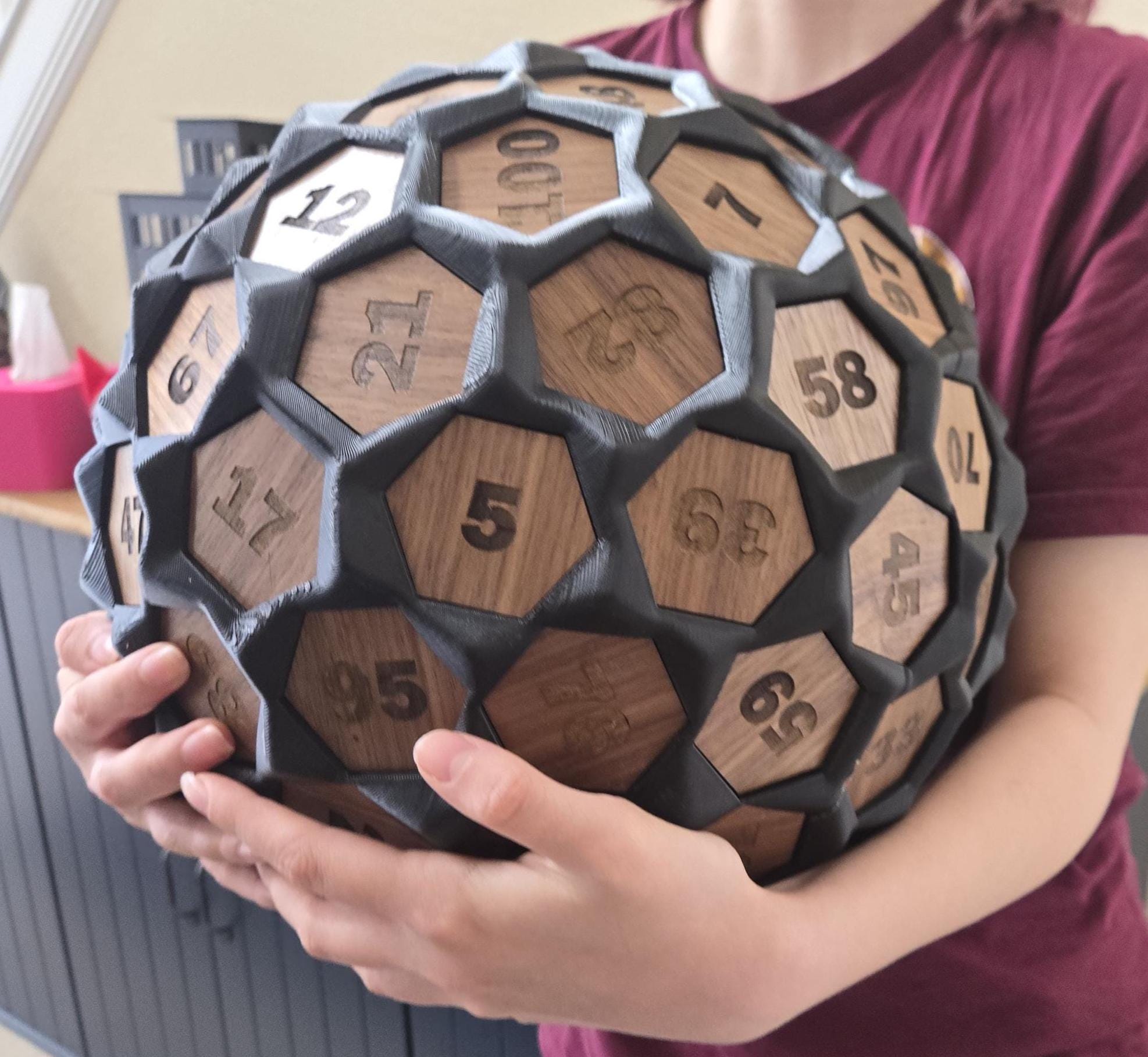A person holding a large, intricate, 3D-printed dice with solid walnut inlay faces, showcasing the numbers 1 through 100.