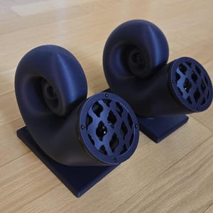 May include: Two navy blue, horn-shaped speakers with a unique, spiral design. Each speaker has a circular opening with a patterned grill. They sit on square bases and are placed on a light wood surface.