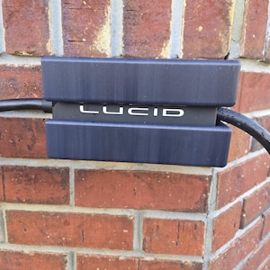 Puede incluir: Un dispositivo rectangular azul oscuro con la palabra "LUCID" en blanco está montado en una pared de ladrillos rojos. Un cable negro sale del dispositivo.