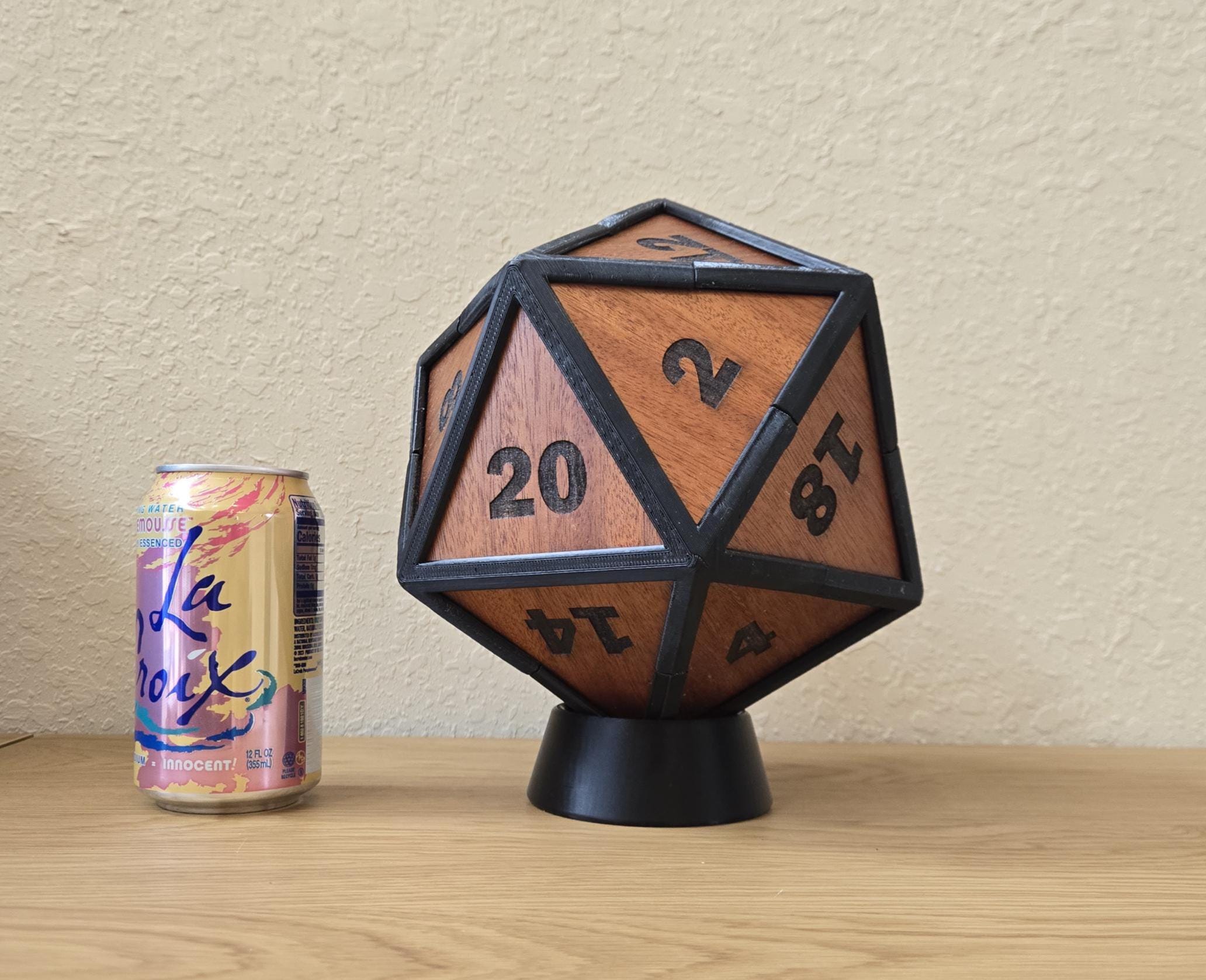 A giant mahogany D20 dice with rubberized edges, standing on a black base, next to a can of LaCroix sparkling water.