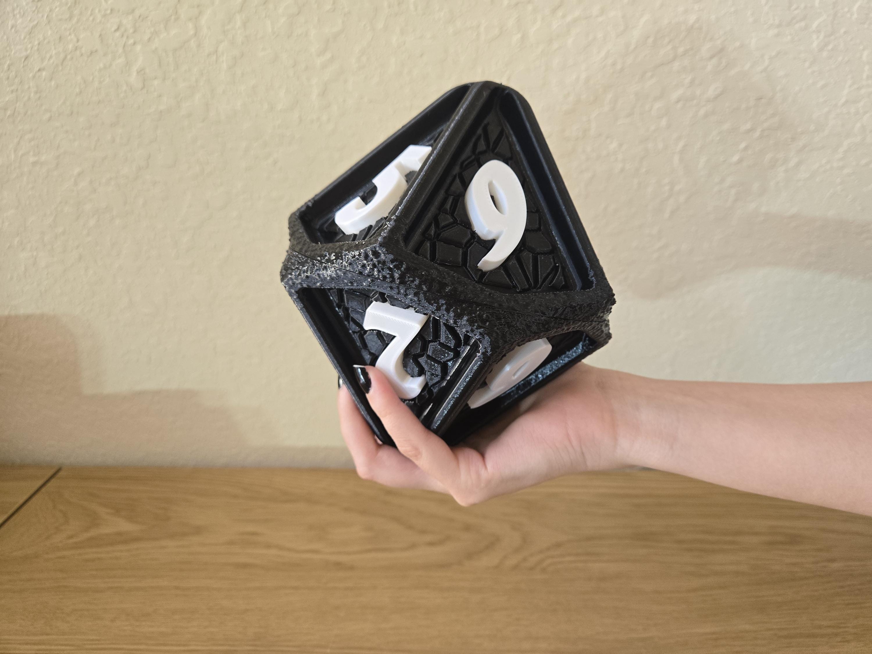 A hand holding a large, black, rubberized D10 die with white numbers on a wooden surface.