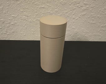 May include: A cylindrical, beige container with a matching lid. The container is made of a matte material and sits upright on a dark surface. The lid fits snugly on top, creating a seamless look. The container is designed to hold small items.