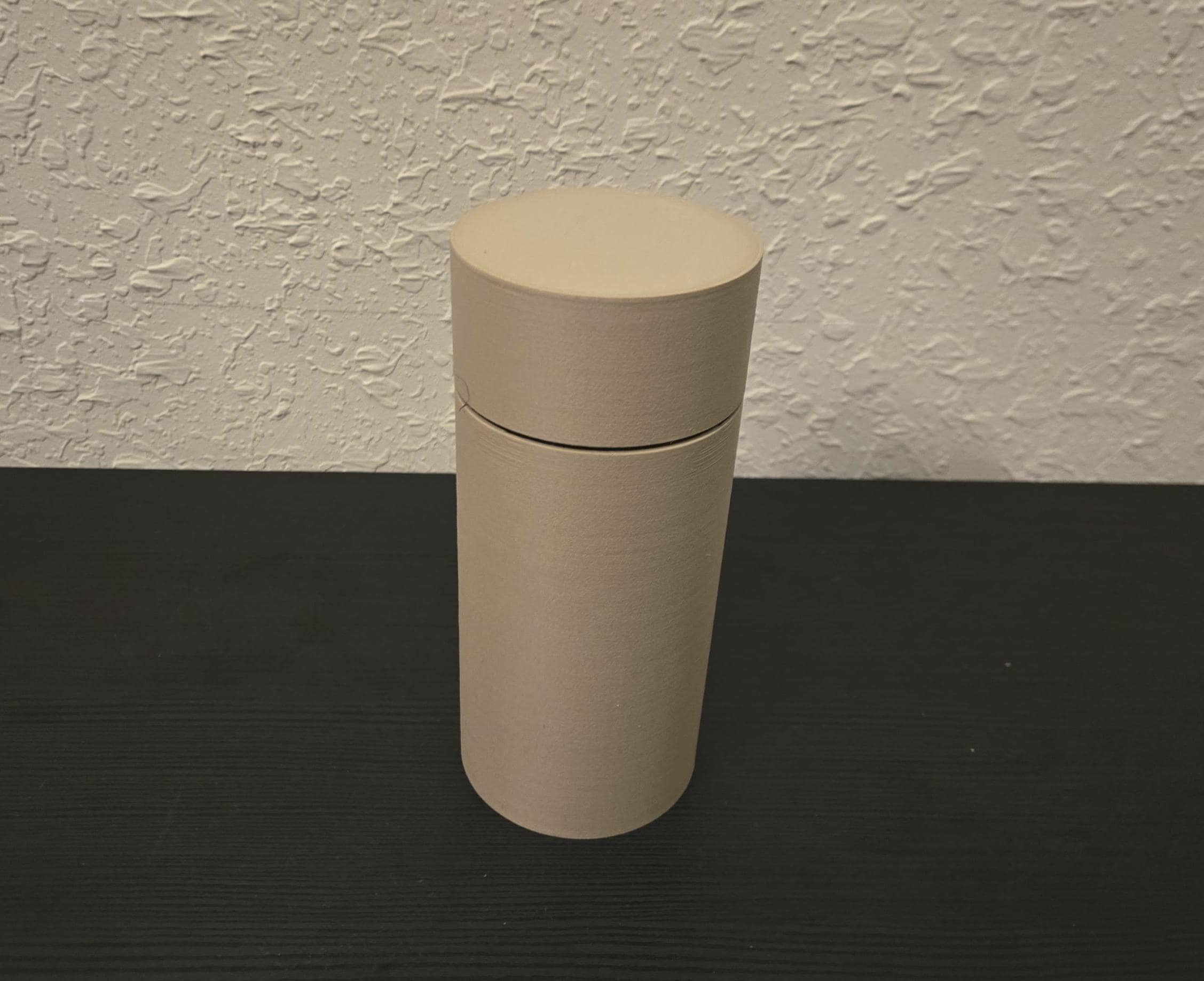 A beige cylindrical cigar holder with a lid, placed on a black surface against a textured wall.