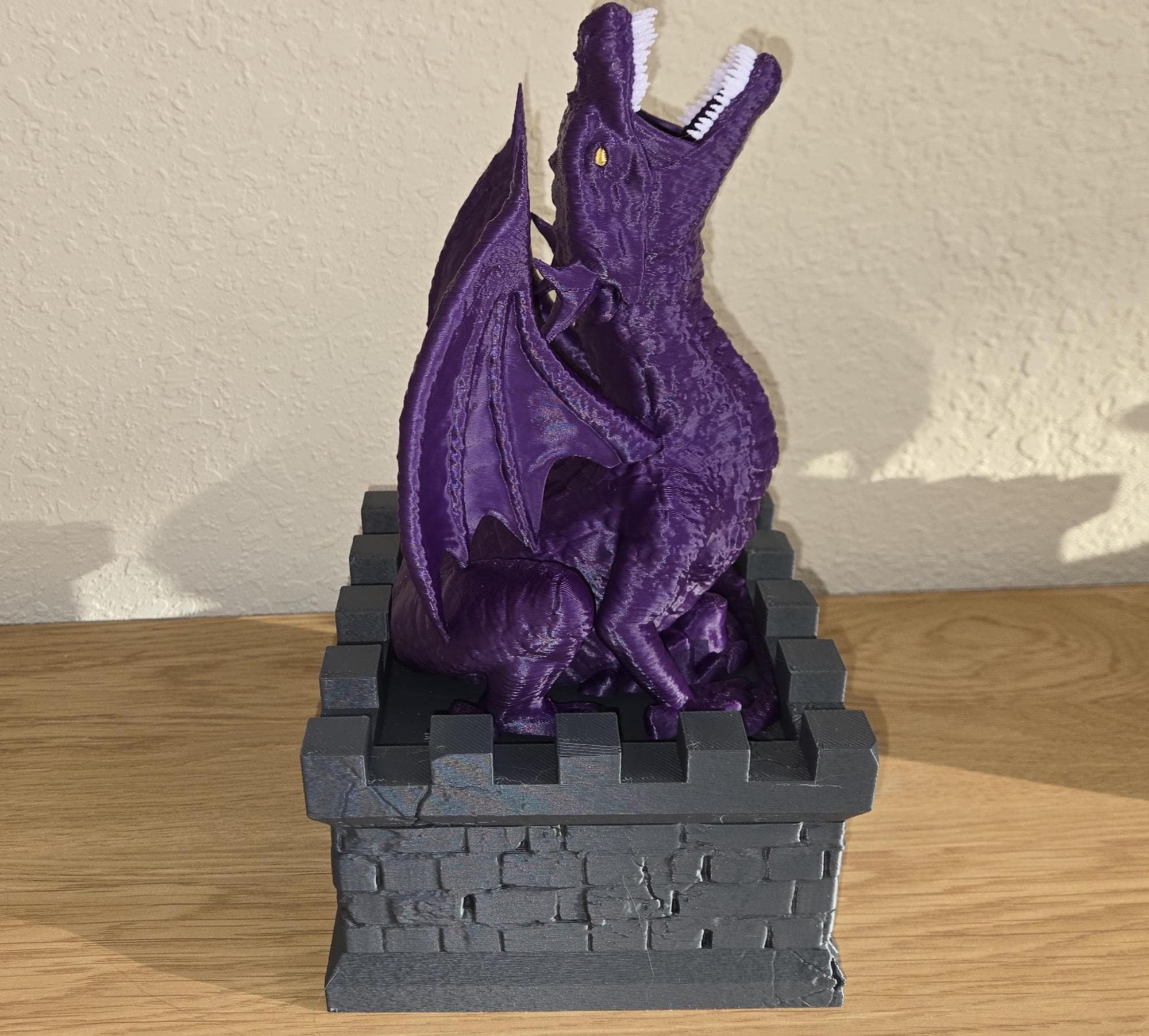 A purple dragon figurine sitting on a black castle wall, with a yellow eye and white teeth, placed on a wooden surface.