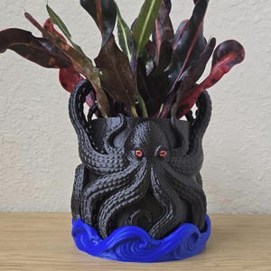 Pode incluir: Vaso preto em forma de polvo com olhos vermelhos e base de ondas azuis, segurando uma planta com folhas vermelhas escuras e verdes. Um item decorativo único para casa ou escritório.