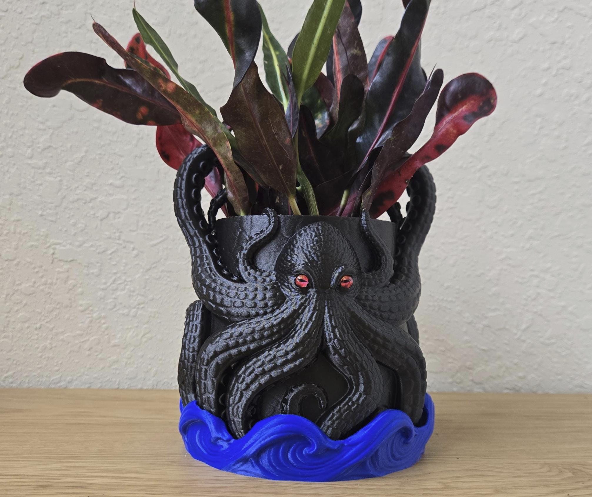 A black octopus planter with red eyes and a blue base, filled with vibrant red and green leaves.