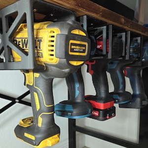 May include: A black metal wall-mounted rack with four power tools hanging from it. The tools are a yellow and black DeWalt drill, a blue and black drill, a black and red drill, and a black and blue drill. The rack is mounted on a wooden surface.