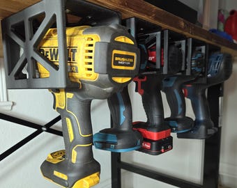 Peut inclure: Une série de quatre perceuses électriques suspendues à un support de rangement en plastique noir imprimé en 3D, fixé sous une étagère en bois. Les perceuses présentent diverses couleurs dont le jaune, le noir, le rouge et le bleu.