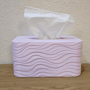 May include: A rectangular, light purple tissue box with a wave-like pattern on the sides. White tissues are pulled from the top opening. The box sits on a wooden surface.