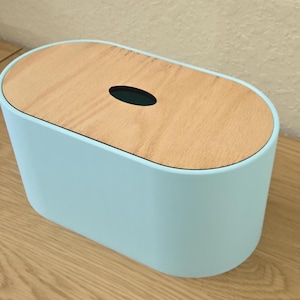 May include: An oval-shaped tissue box cover with a light blue base and a flat, light-toned wooden lid. The lid features a centered, elongated oval opening for dispensing tissues, providing a clean and functional design for home or office use.