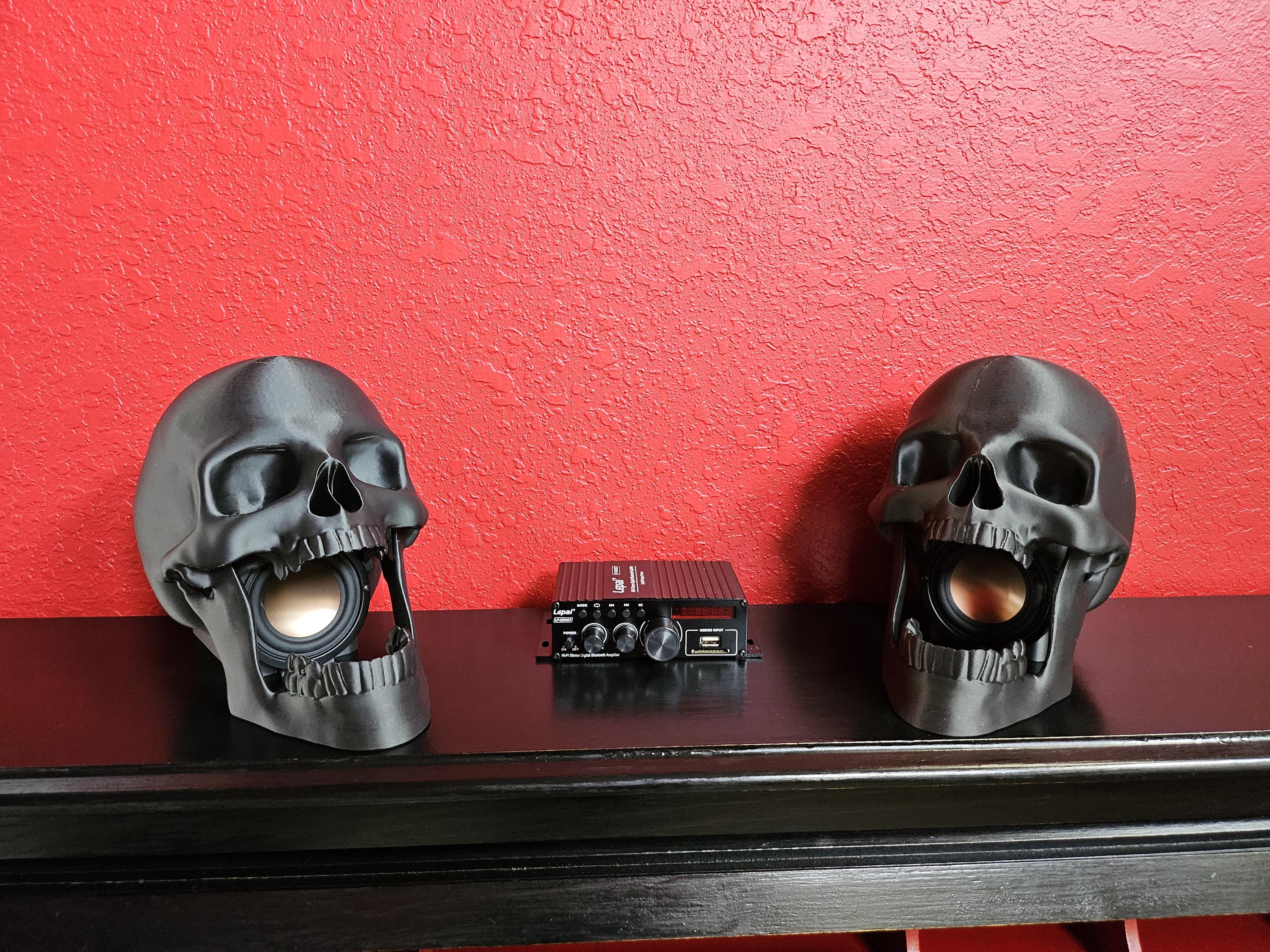 A pair of black skull speakers with gold-colored speakers, placed on a black shelf against a red textured wall, with a small.