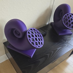 May include: Two purple, horn-shaped speakers with a lattice-style speaker cover. The speakers are on a black wooden surface. The speakers have a unique, artistic design, and are likely used for audio playback.