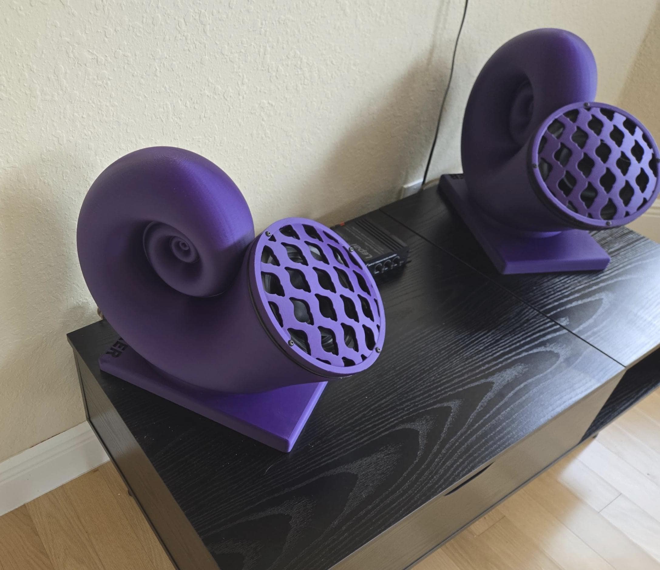 A pair of large, purple nautilus speakers with a grid pattern on the front, placed on a black wooden surface.