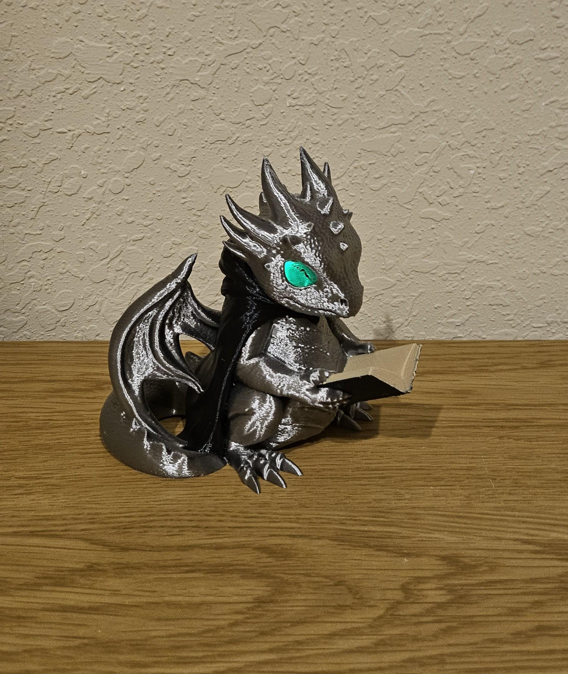 A glowing dragon figurine holding a book, placed on a wooden surface against a textured wall.