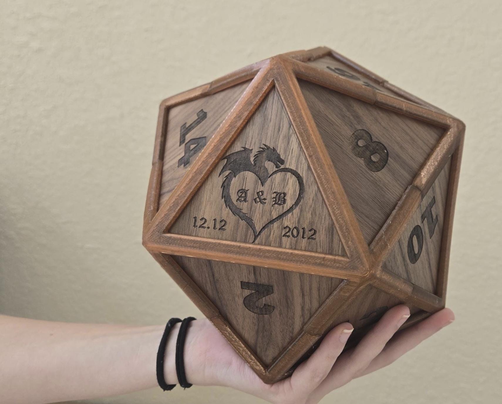 A hand holding a large, wooden, d20-shaped object with personalized engravings, including a heart, initials, and a date.