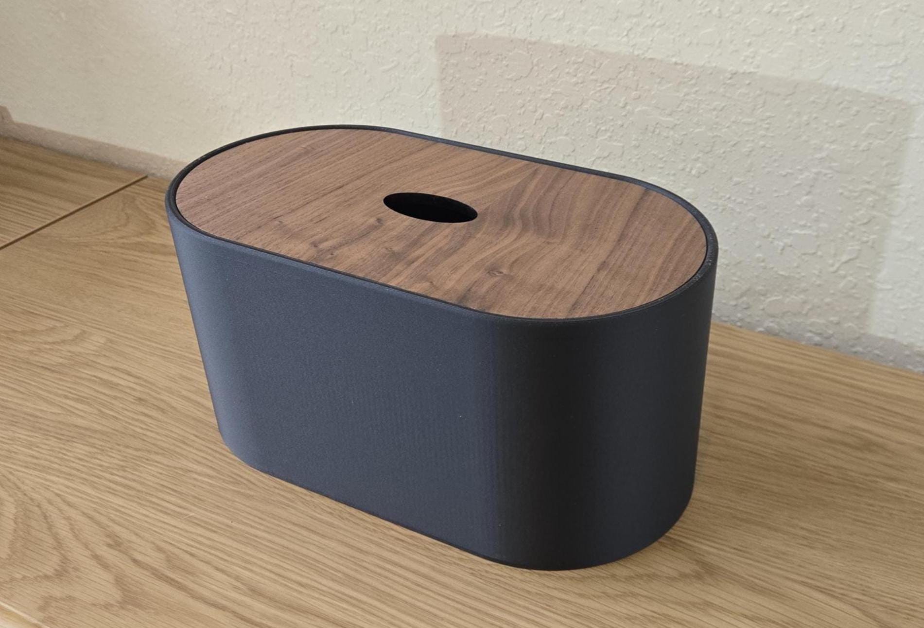 A modern, oval-shaped toilet paper roll storage box with a carbon black exterior and a walnut wood lid, placed on a wooden表面.