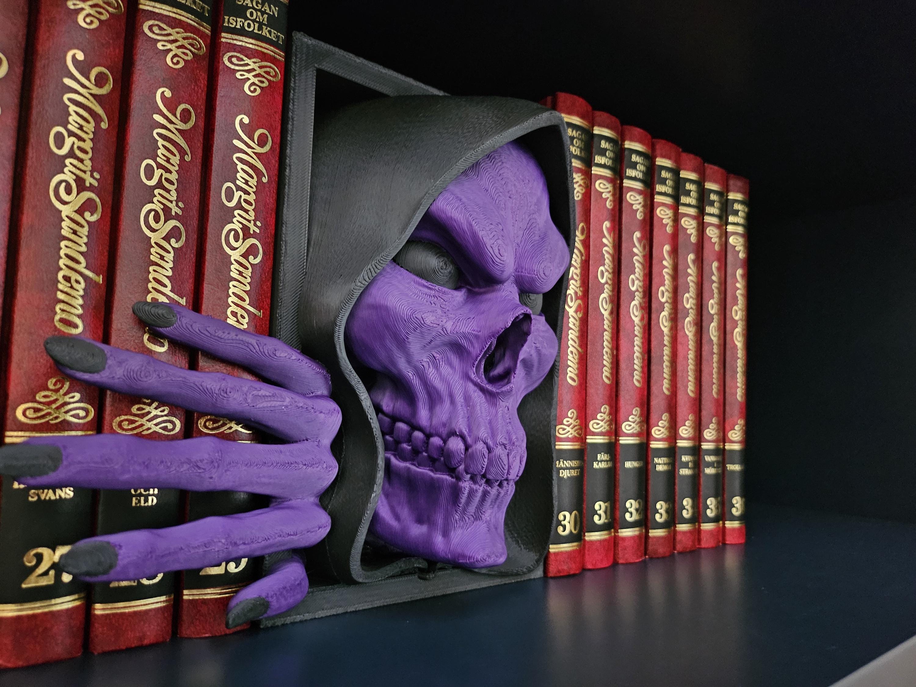 A black and purple skull-shaped bookshelf insert with a hand reaching out from the side, placed on a shelf with red books.