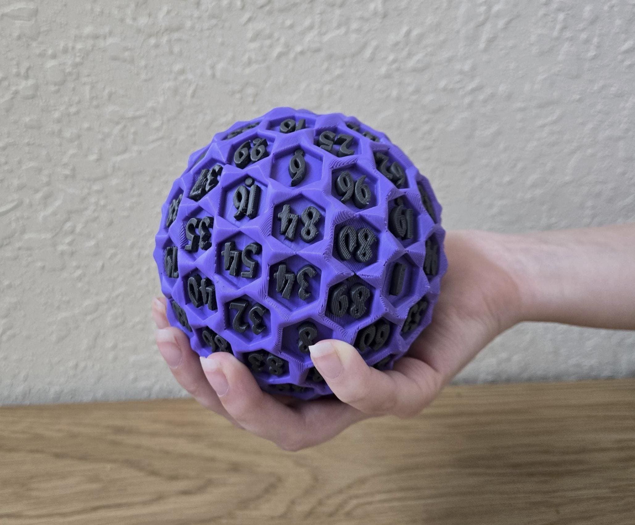 A hand holding a purple, 3D-printed, honeycomb-patterned ball with numbers on it, likely a giant percentile die for role-play.