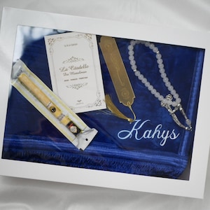 Peut inclure: Présentation encadrée avec un cigare dans un emballage, un signet doré, un chapelet de perles blanches et une carte avec du texte en français. Les articles reposent sur un tissu bleu, avec le nom "Kahys" brodé en blanc.