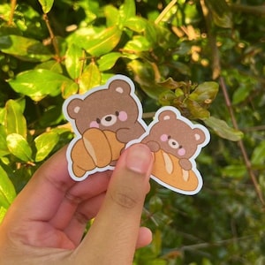 May include: Two die-cut stickers featuring a brown bear character with pink cheeks holding a golden-brown croissant. The stickers have a white border and a matte finish, showcasing a cartoon illustration of a bear hugging a pastry.
