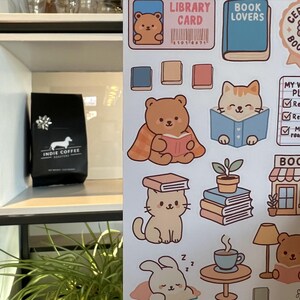 Puede incluir: Una bolsa de caf&eacute; negra con el texto "INDIE COFFEE ROASTERS" y una ilustraci&oacute;n de perro salchicha est&aacute; sobre una estanter&iacute;a blanca. Junto a ella, pegatinas con ilustraciones tem&aacute;ticas de libros, incluyendo osos, gatos, libros y las palabras "LIBRARY CARD" y "BOOK LOVERS".