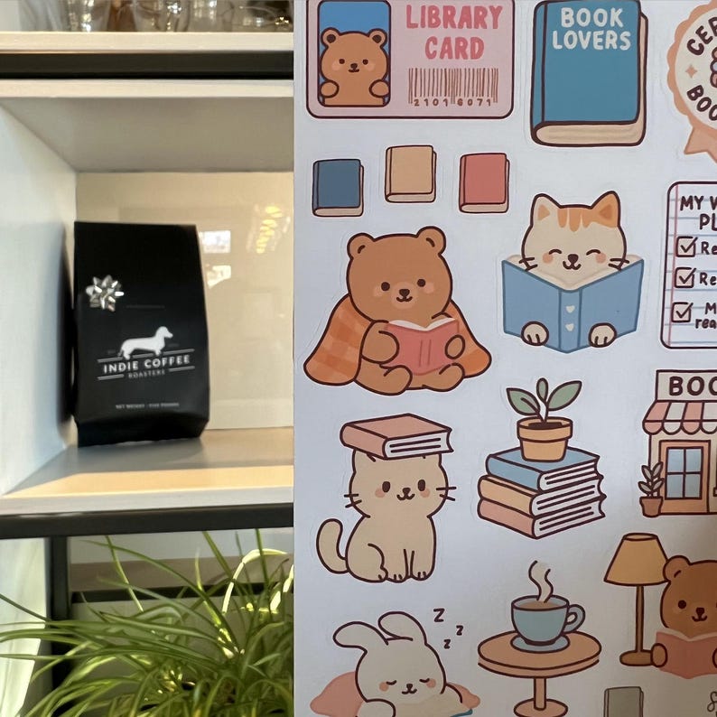 Puede incluir: Una bolsa de caf&eacute; negra con el texto "INDIE COFFEE ROASTERS" y una ilustraci&oacute;n de perro salchicha est&aacute; sobre una estanter&iacute;a blanca. Junto a ella, pegatinas con ilustraciones tem&aacute;ticas de libros, incluyendo osos, gatos, libros y las palabras "LIBRARY CARD" y "BOOK LOVERS".