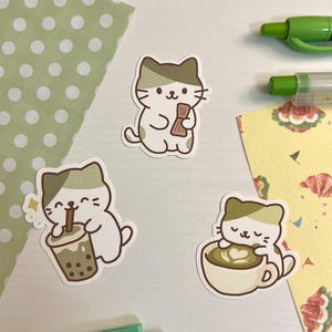 May include: Three cat stickers with a white background and a green and brown color scheme. One cat holds a bundle, another drinks boba tea, and the third is in a coffee cup. The stickers are on a white surface with green and yellow paper.