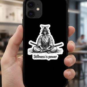 以下が含まれることがあります： 黒い携帯電話ケース。瞑想する侍の白黒イラストで、後ろに2本の剣が交差しています。「Stillness is power」の文字が下に表示されています。